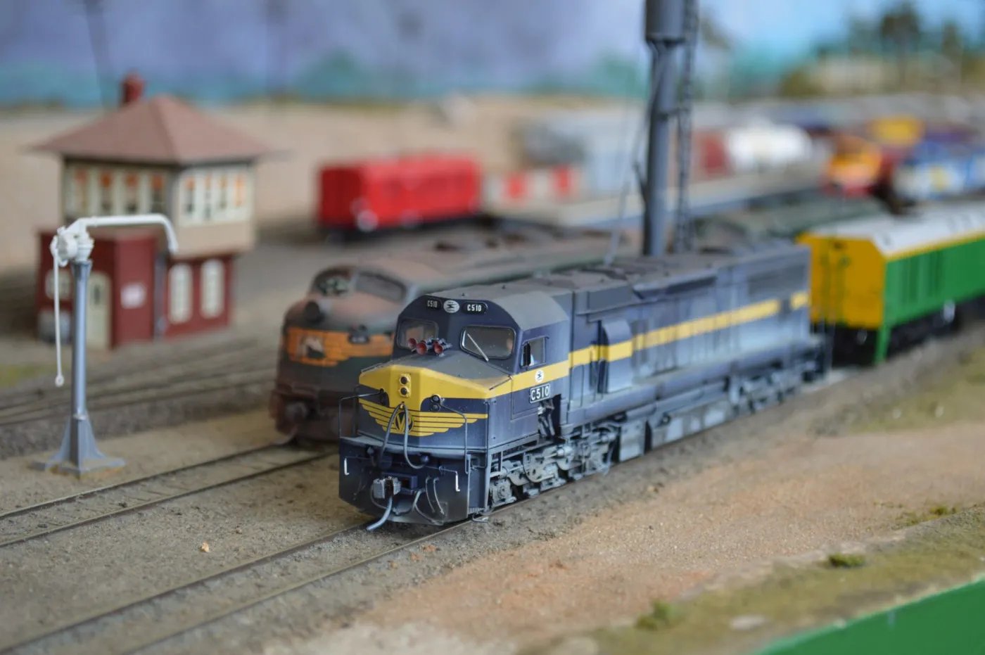 A dark blue and yellow locomotive numbered 2310 pulls a green passenger car on a model train track, with a red building and signal post in the background.