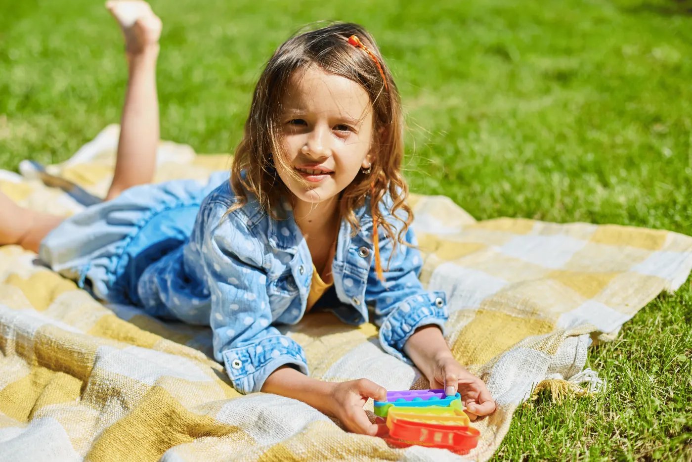 A young girl with long brown hair lies on a yellow and white checkered blanket on a grassy lawn. She wears a blue denim shirt and holds a colorful toy with red, yellow, blue, and