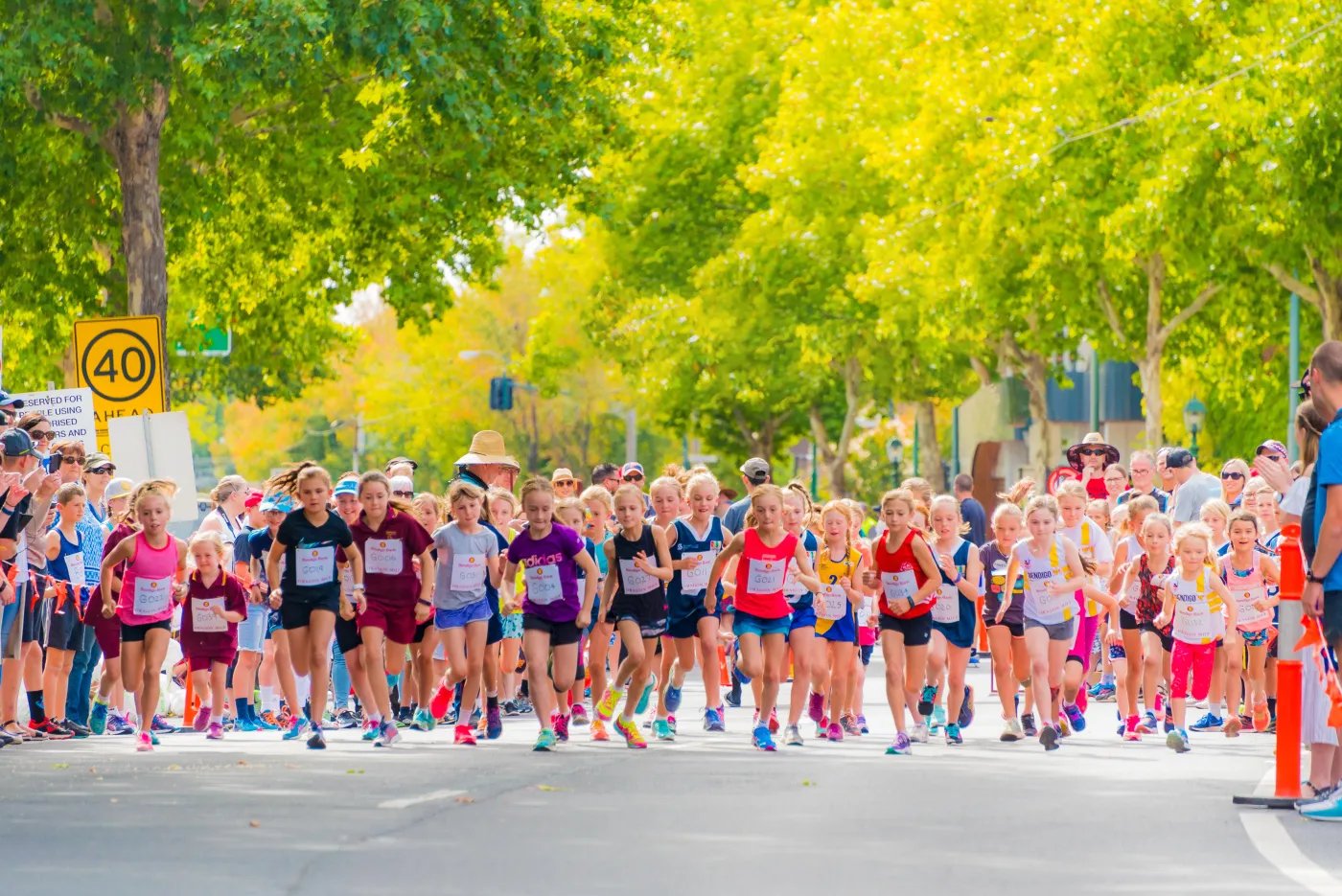 A large group of runners in colorful athletic clothing participate in a race on a tree-lined street, with a speed limit sign visible on the left.
