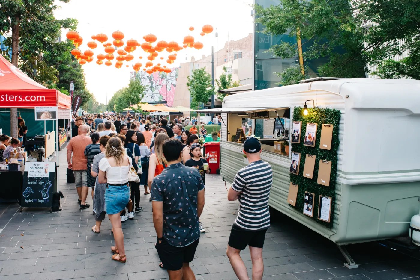 A white food truck with green trim and hanging greenery stands out among a crowd of people at an outdoor market, with orange balloons floating overhead.