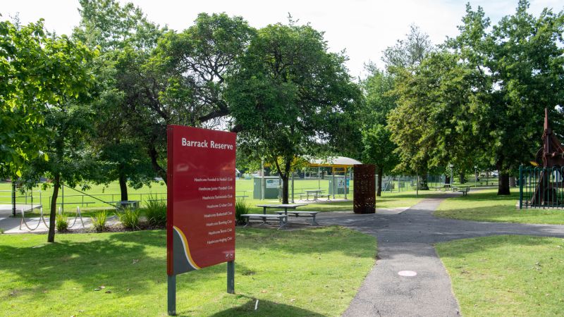 A red sign labeled "Barrack Reserve" stands on grass near a paved path, with trees and a playground visible in the background.