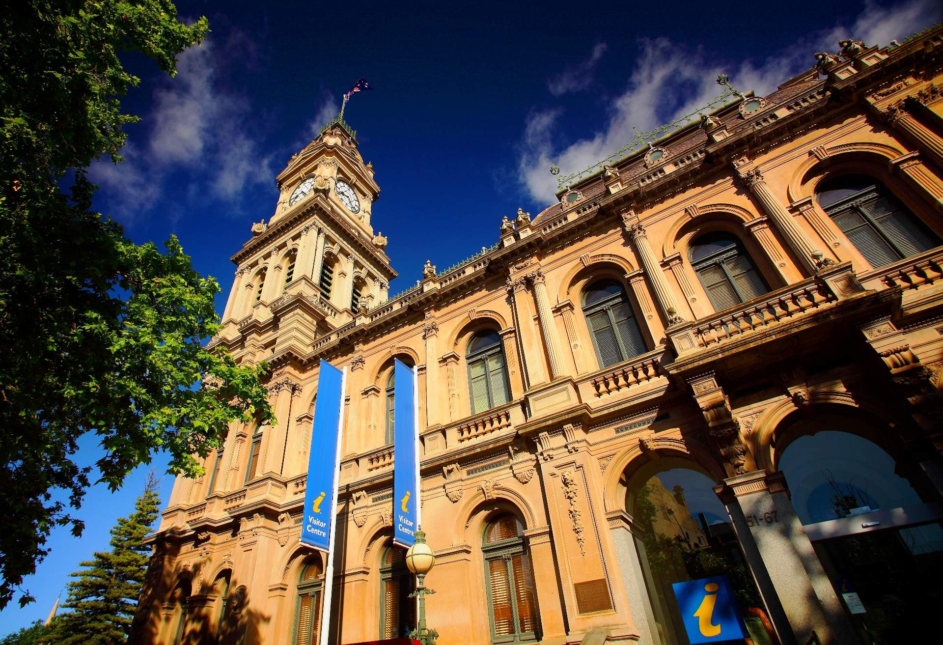 A tan stone building with arched windows and a clock tower topped by a flag stands under a blue sky with clouds, flanked by trees and a street lamp.
