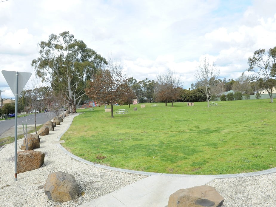 A grassy park with trees and a sidewalk bordered by rocks, with a road and houses visible in the distance.