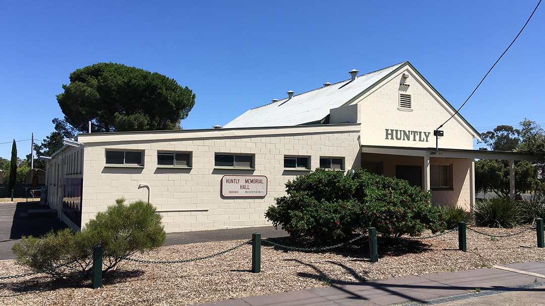A white single-story building with a sloped roof and the word "HUNTLY" on its facade, identified as the Huntly Memorial Hall, is situated on a street with bushes, gravel, and a sidewalk
