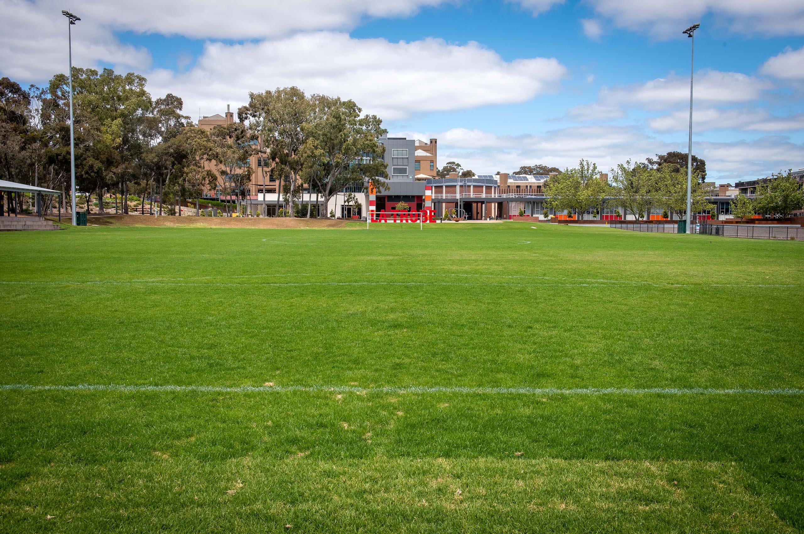 A grassy sports field with white boundary lines, flanked by trees and a building in the background, under a blue sky with clouds.