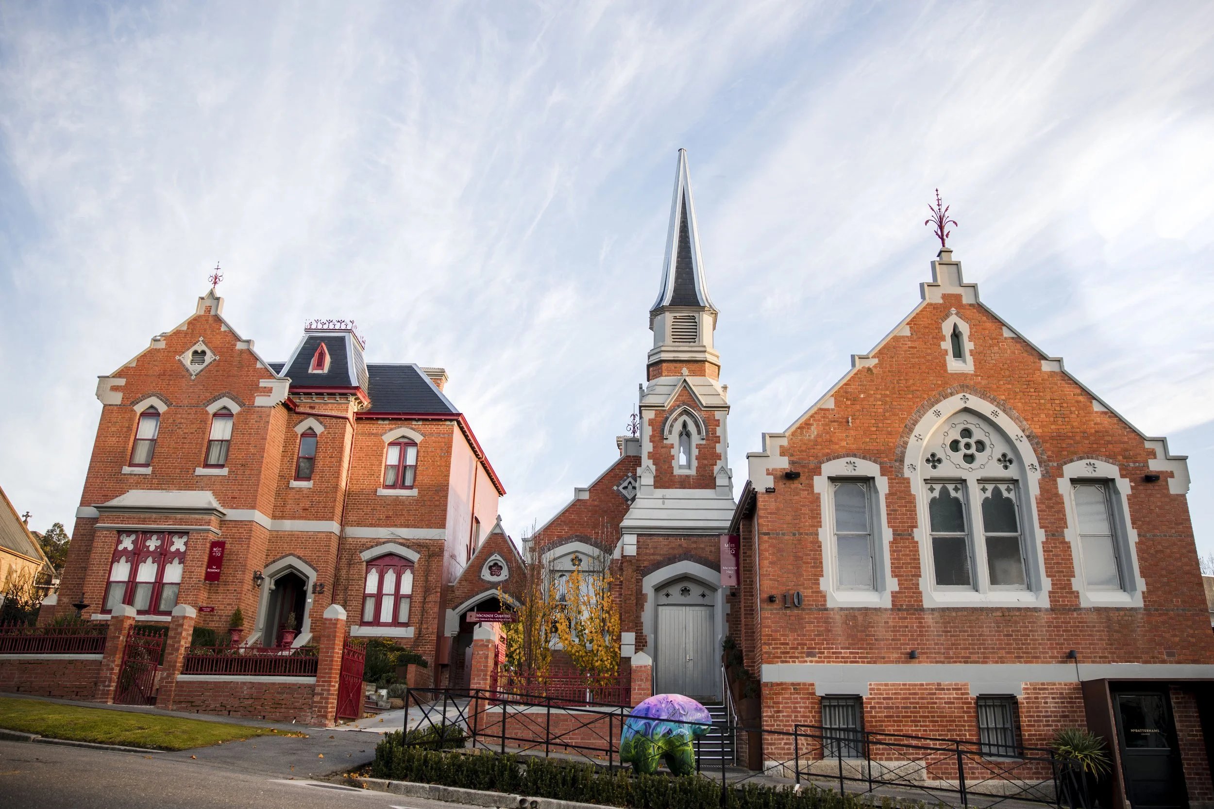 A red brick church with a tall steeple and cross stands beside a smaller red brick building under a partly cloudy sky, with a purple umbrella and fence in the foreground.