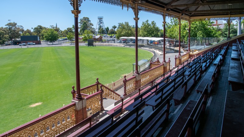 A grand stand with ornate red and gold railings overlooks a lush green field with trees and a distant tower.
