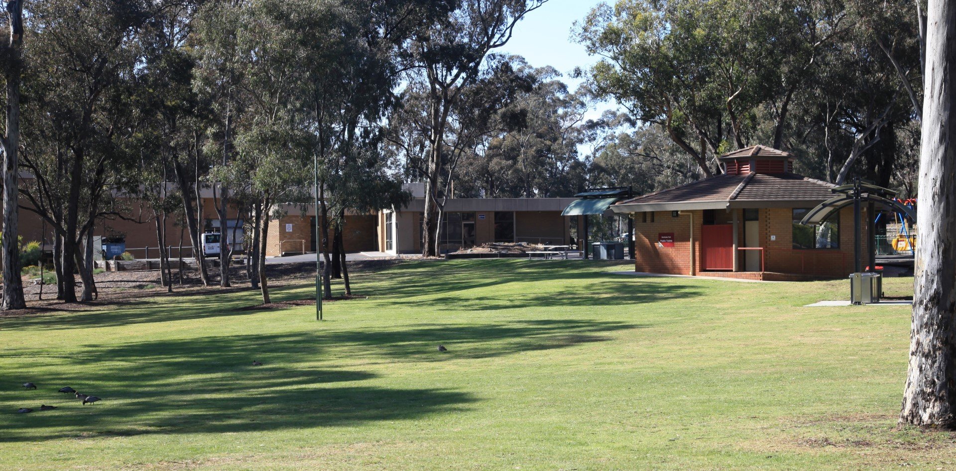 A grassy area with trees, a small brown building with a red door, and a picnic shelter is pictured under a clear blue sky.