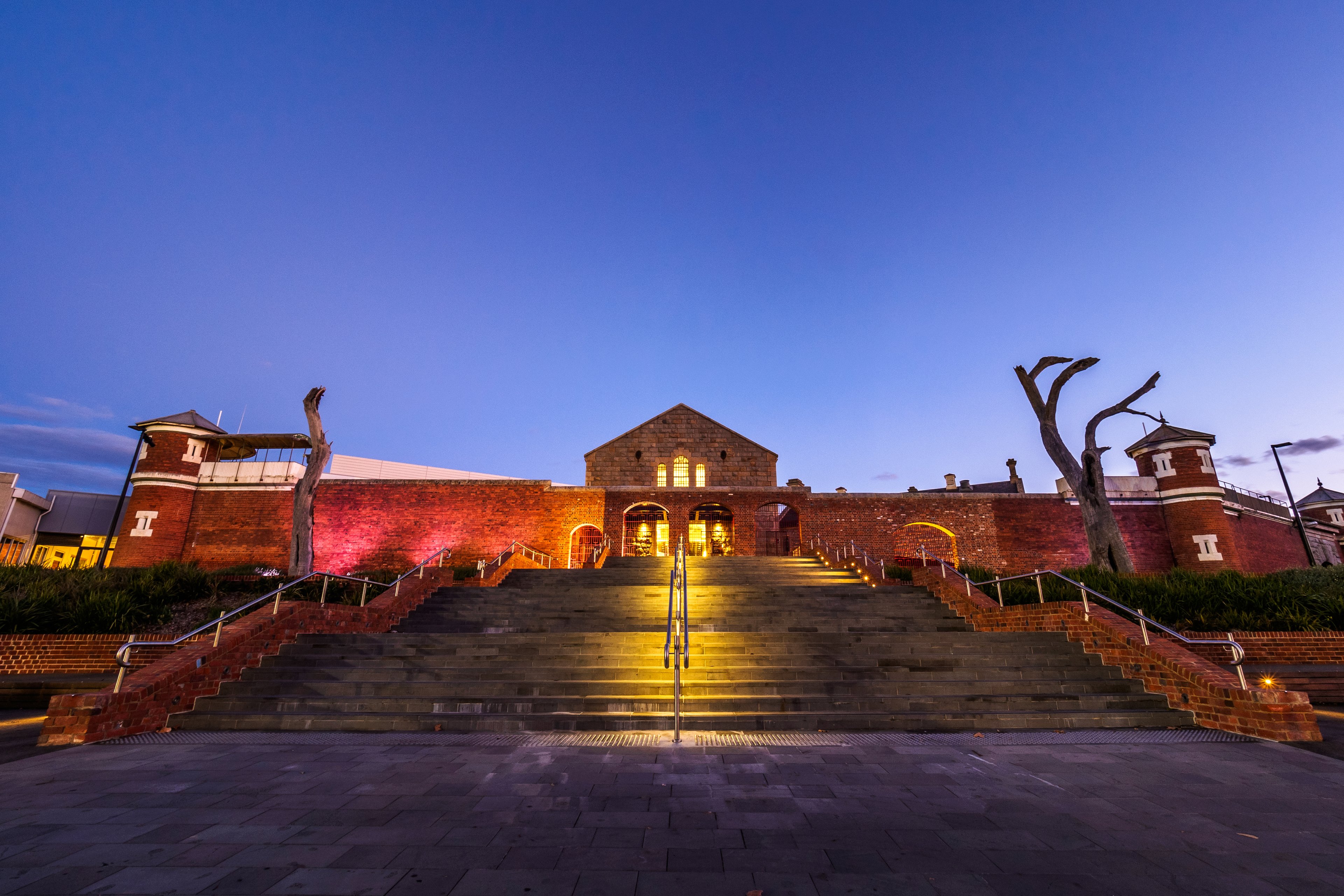 A brick building with arched windows and a central entrance is illuminated at dusk, flanked by brick walls and stairs, with trees on either side.
