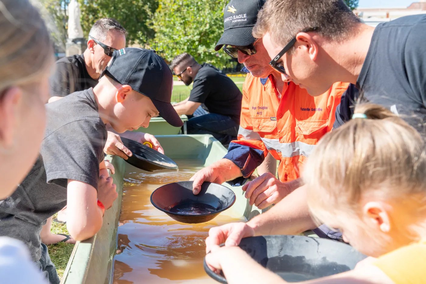 Children and adults gather around a green trough filled with water; some hold black bowls.