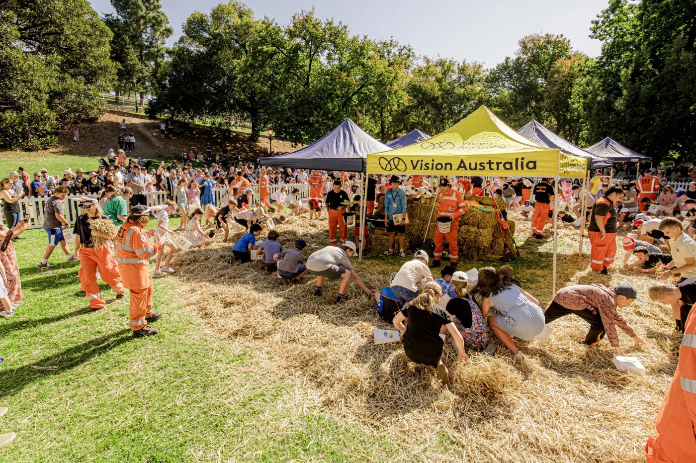 A group of people in orange safety vests and hard hats are working together on a grassy area, with several tents and trees in the background.
