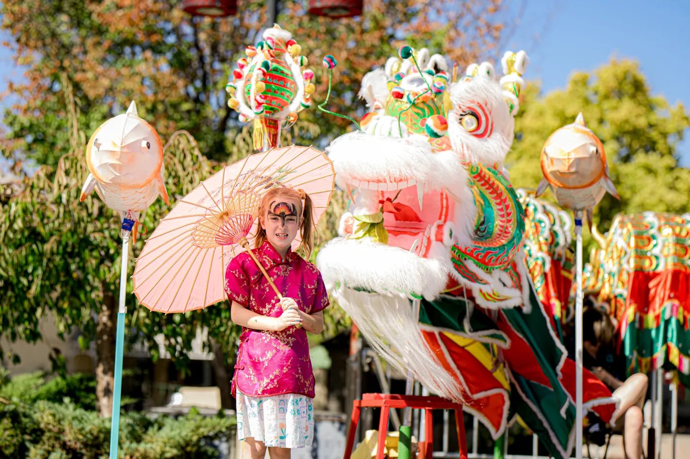 A person in a pink dress holds a pink parasol beside a large, colorful dragon float with elaborate decorations and additional floats in the background.
