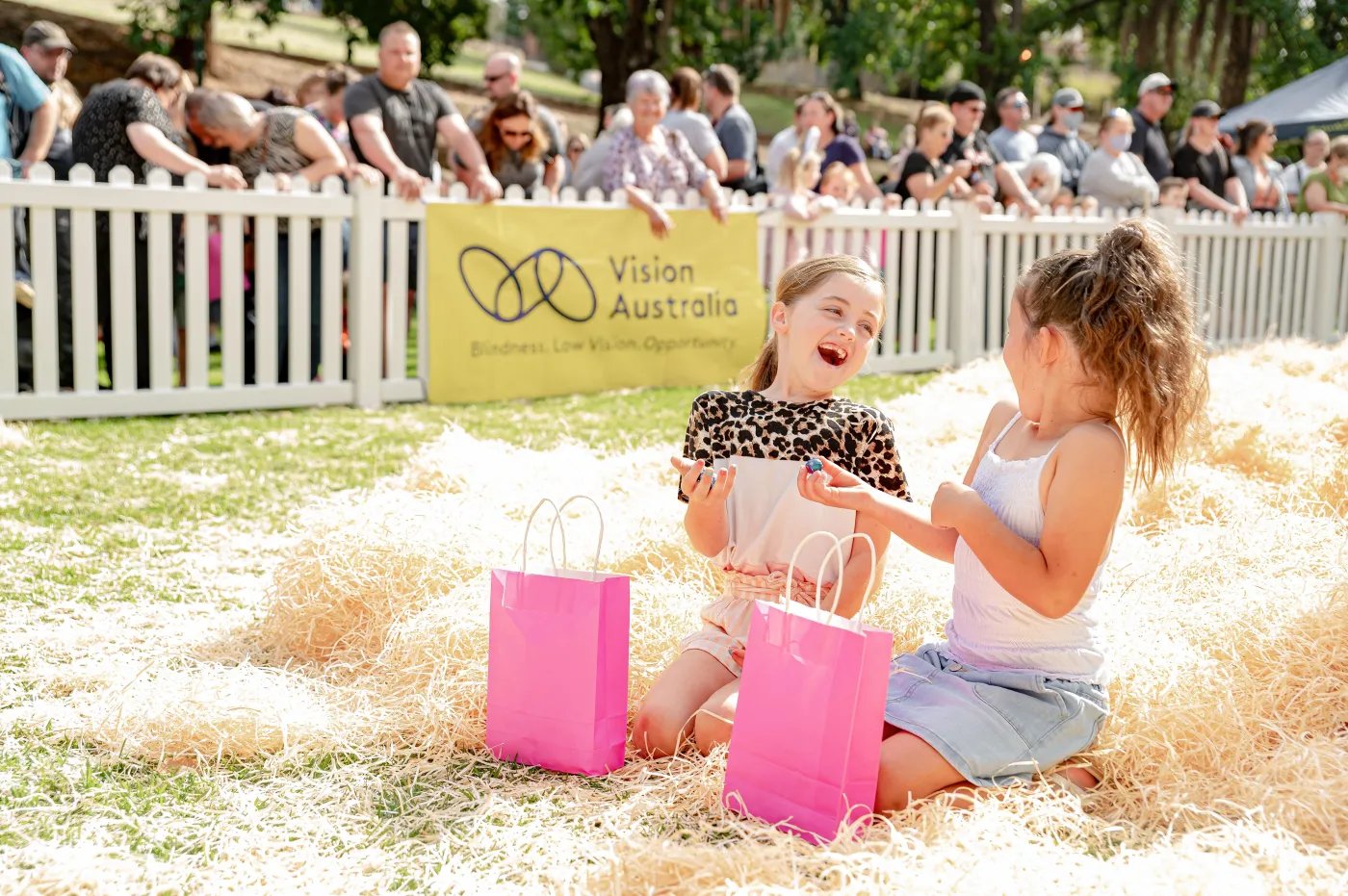 Two girls sit on hay near pink and pinkish-purple bags, with a white picket fence and a yellow banner reading “Vision Australia” behind them, with spectators visible
