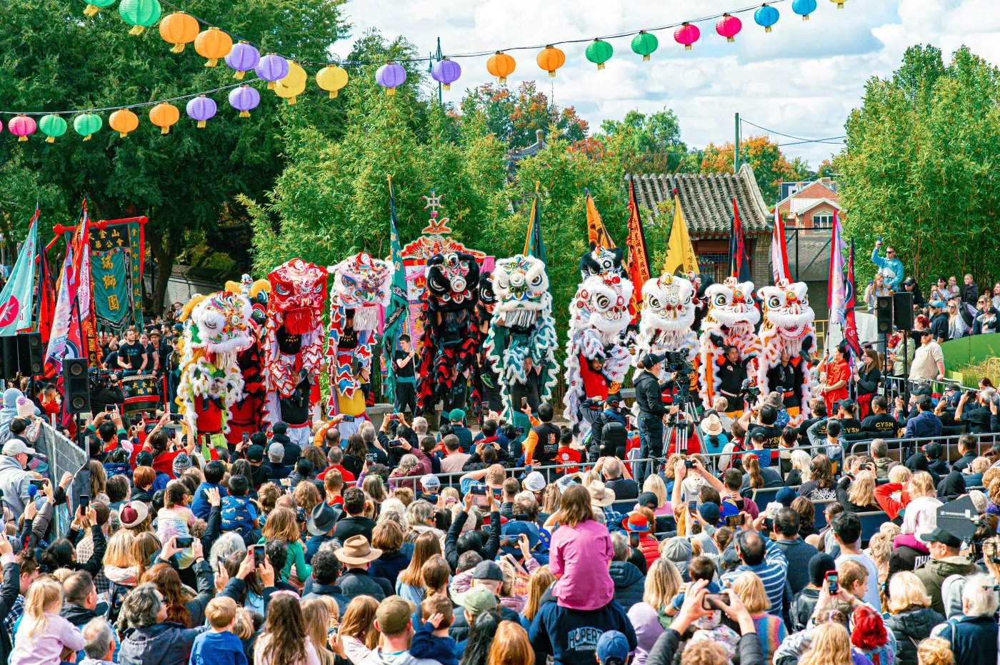 A large crowd gathers outdoors to watch a parade featuring colorful, lion-like costumes adorned with flags and colorful balls, set against a backdrop of trees and festive lights.