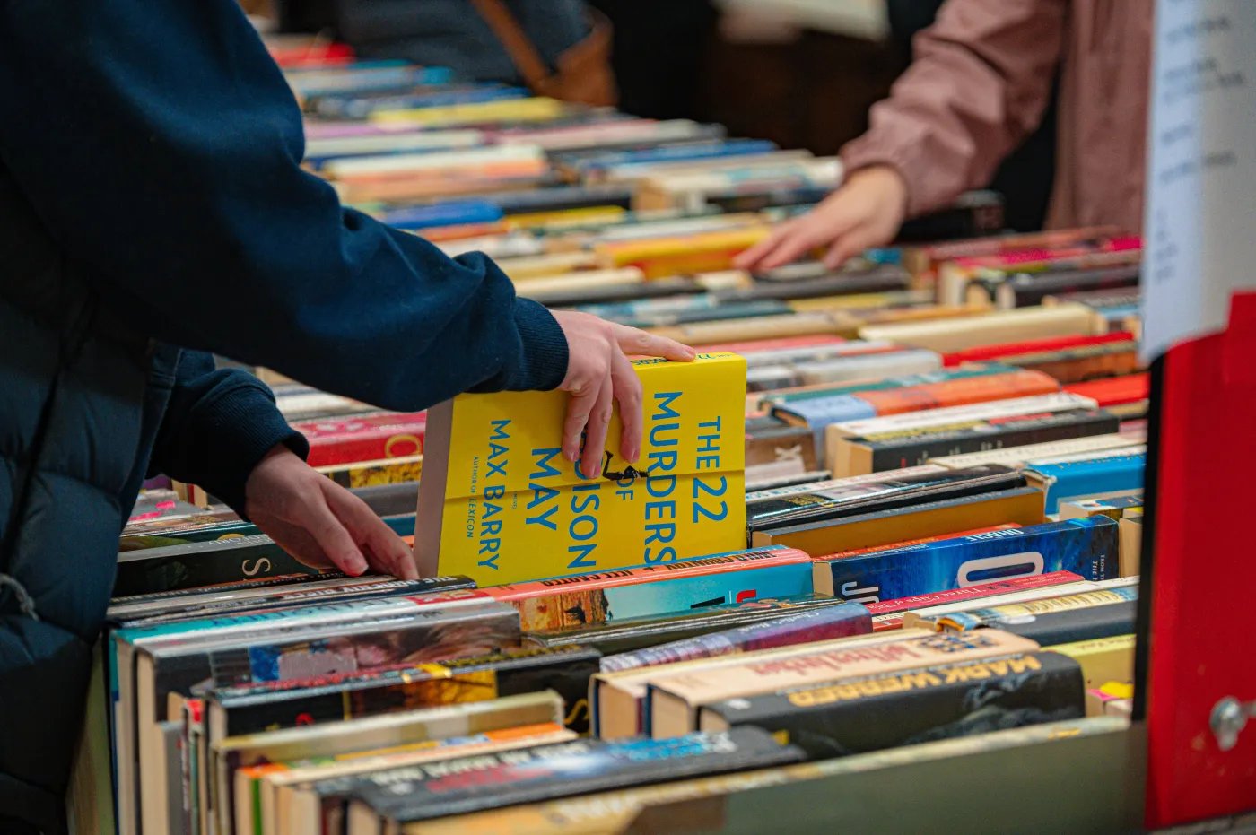 A person in a dark blue jacket reaches for a yellow book titled "The 22 Murders of May" by Max Barry among a diverse collection of books.