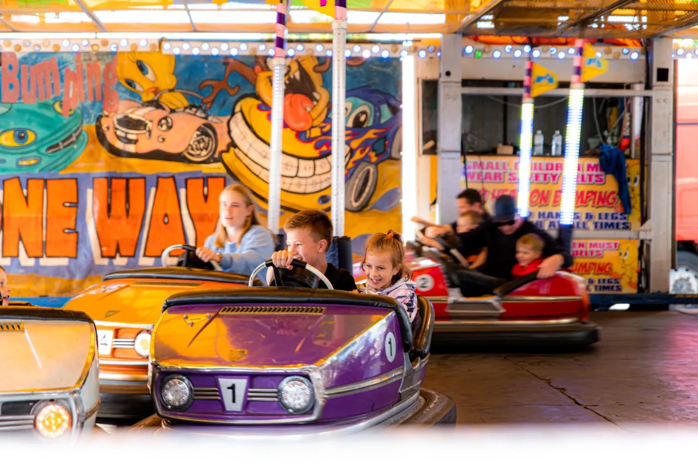 Three children ride bumper cars on a carousel, with a colorful backdrop featuring cartoon characters.