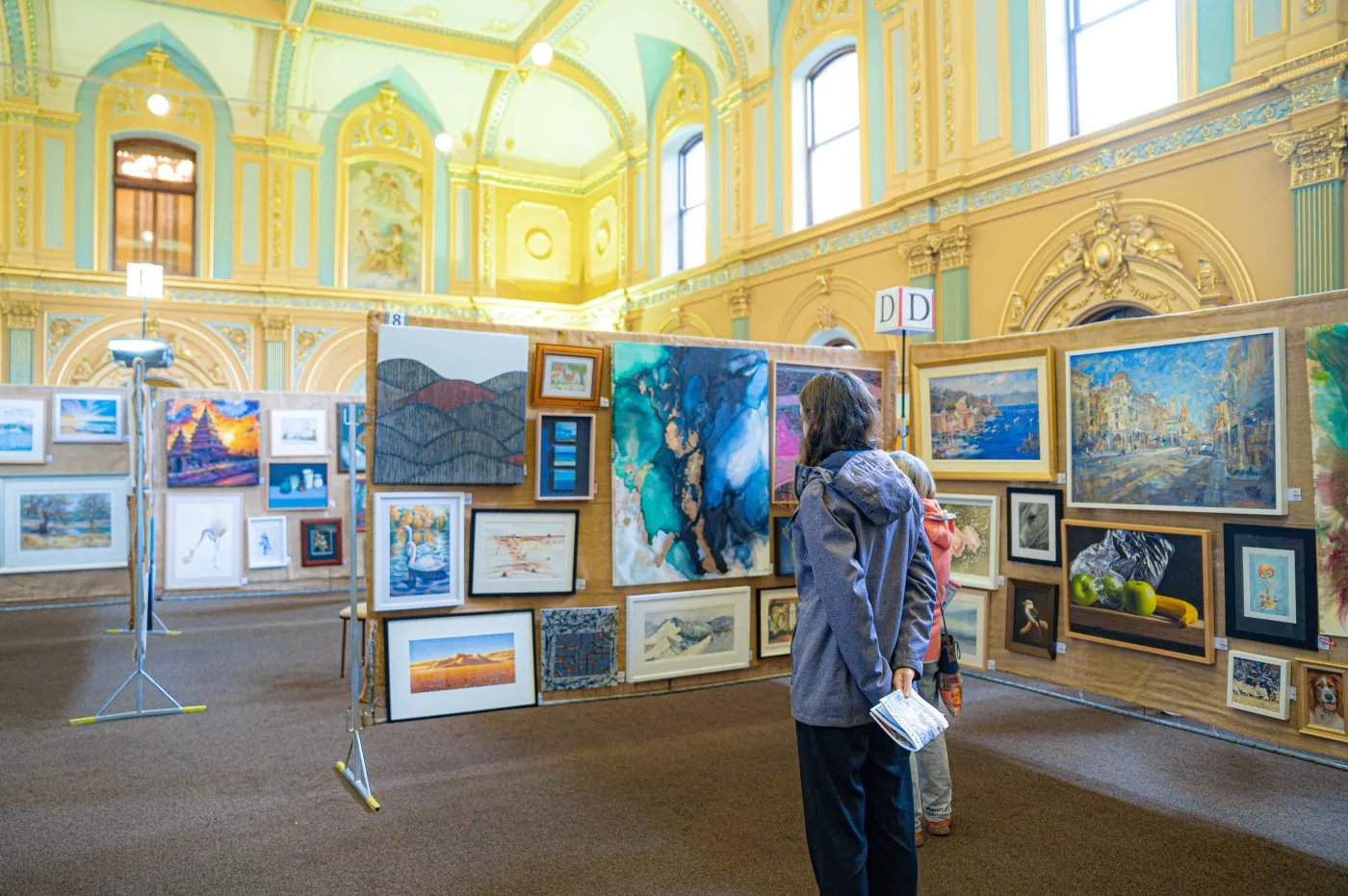 A woman in a gray jacket and dark pants stands near a display of paintings in a large, ornate room with light yellow walls and arched windows.