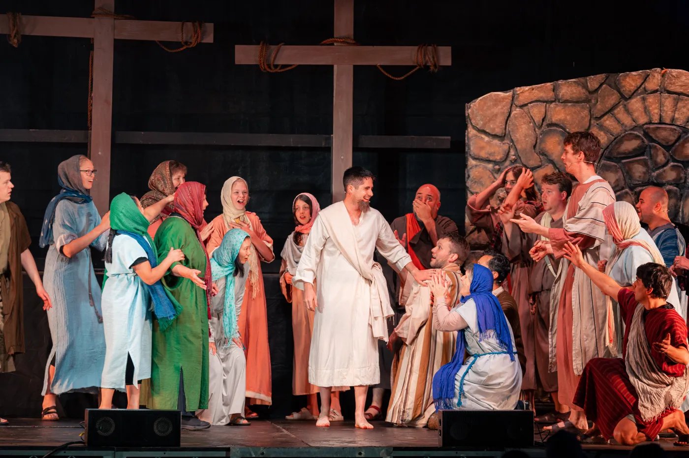 A stage performance features a central figure in a white robe surrounded by people in various costumes, with a stone wall backdrop and wooden cross.