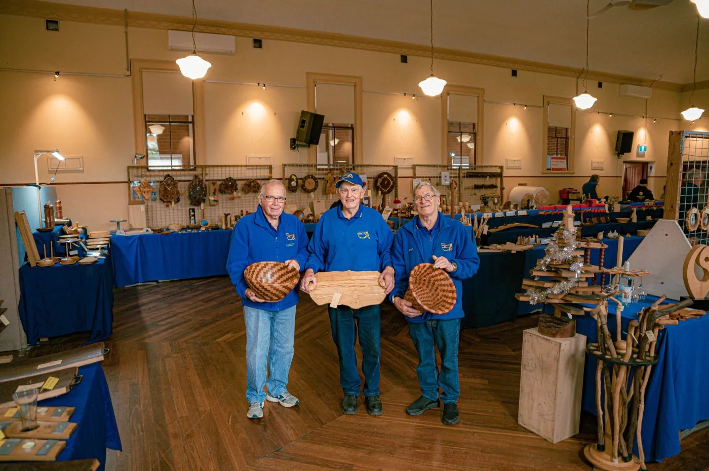 Three men in blue shirts and hats stand in a room with wooden floors and tables displaying various items, including baskets and wooden objects.