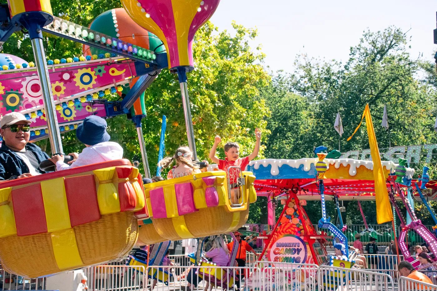 Three children ride a colorful carousel with yellow, red, and pink striped seats under a blue sky with trees in the background.