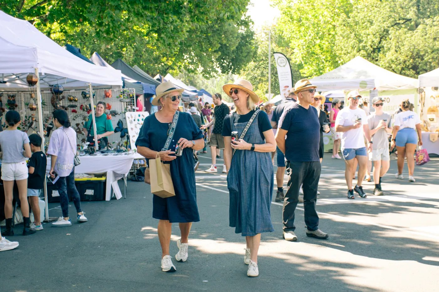 Two women in blue dresses and straw hats walk together, holding drinks, with white tents and trees in the background.