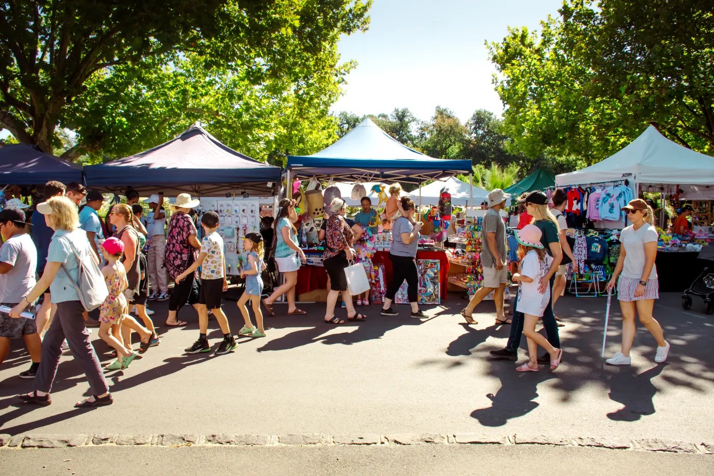A paved street market with multiple tents and stalls under blue and white canopies, with people browsing and walking around. Trees line the street in the background.