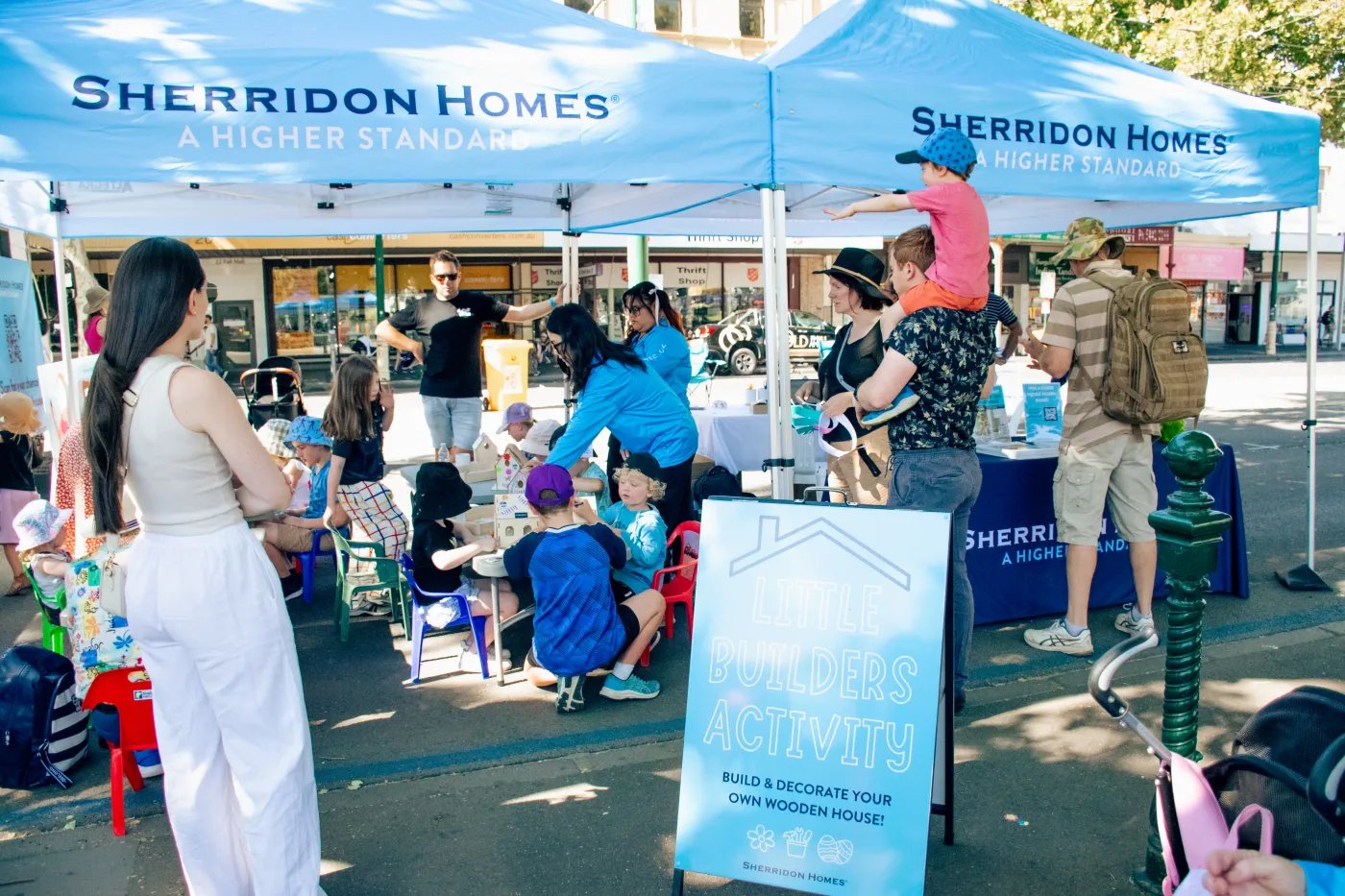 Two blue tents with Sherridon Homes branding are set up in a paved area, with adults and children interacting near tables and a sign