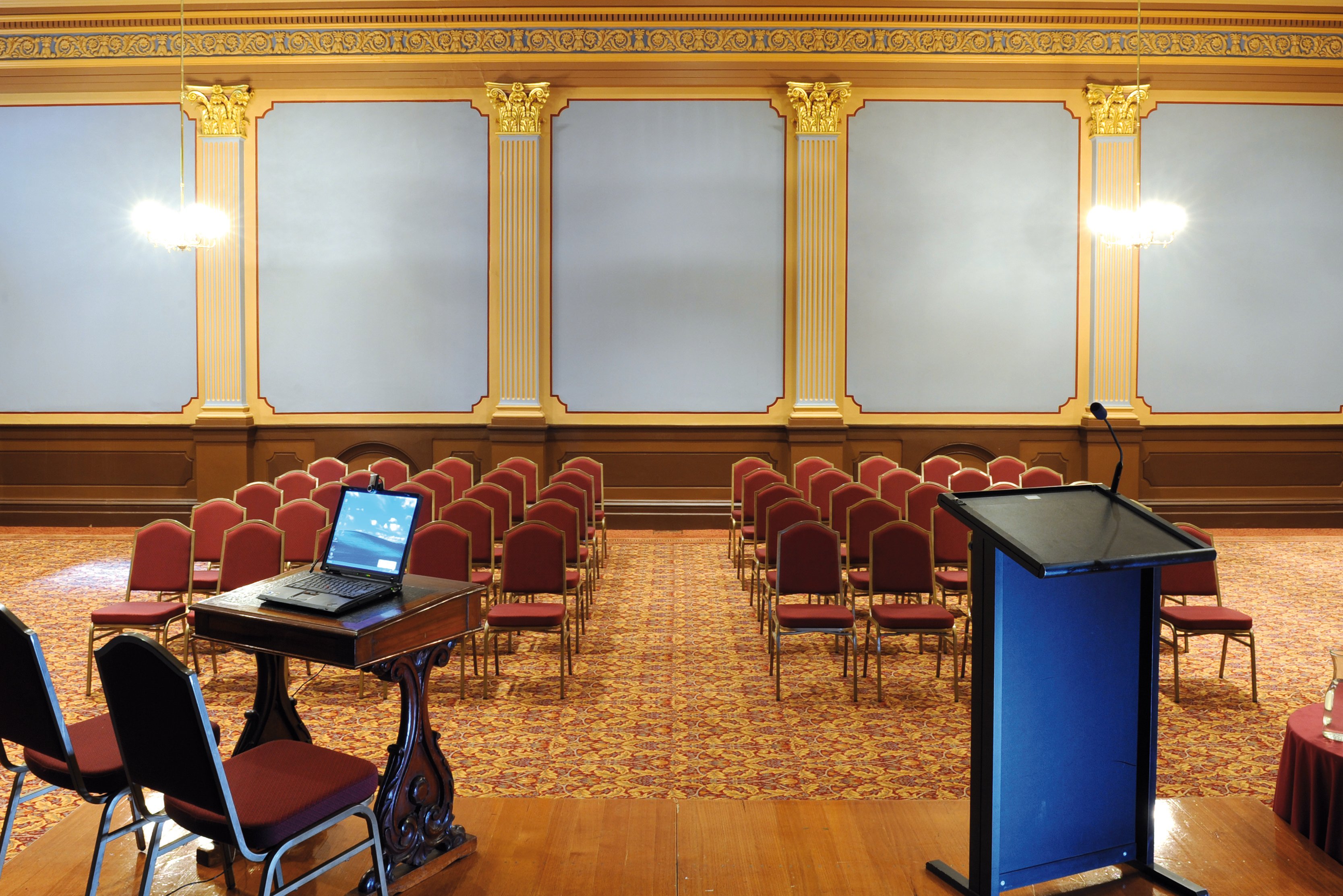 A blue podium with a black top is positioned near a laptop on a table, with rows of red chairs arranged facing the front of a room with light-colored walls and ornate molding.