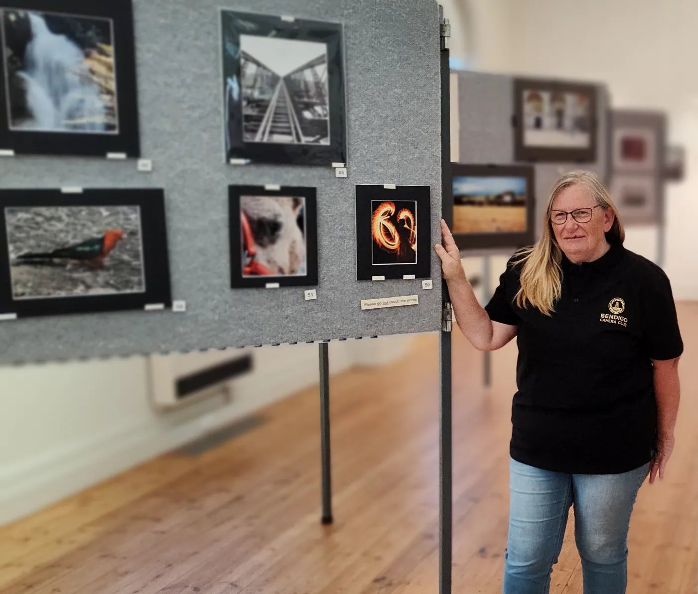 A woman in a black shirt and jeans stands next to a gray bulletin board displaying six photographs of varying subjects. She points toward one of the photos with her right hand. The setting appears to be a gallery or
