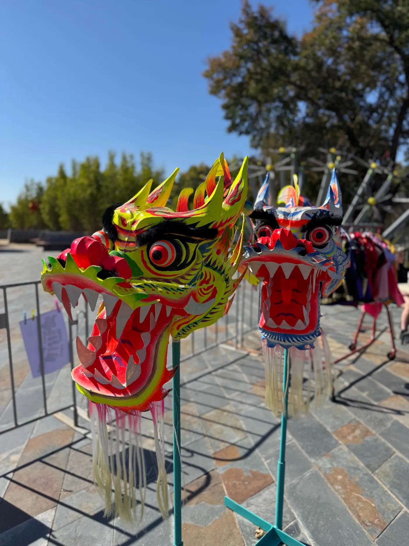 Two brightly colored dragon heads on metal stands stand out against a clear blue sky and stone pavement.