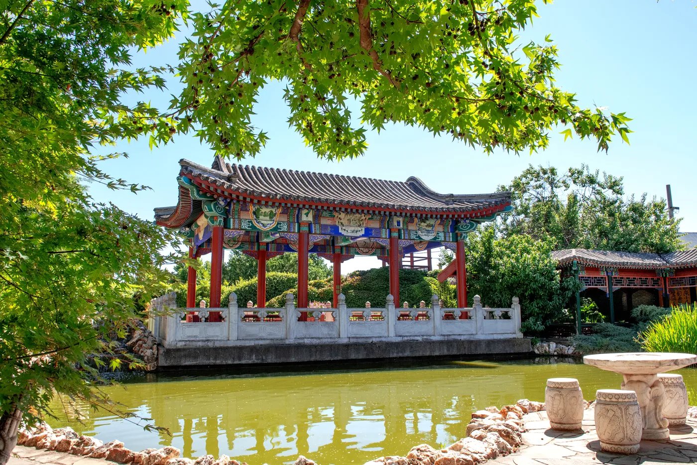 A red-roofed pavilion with white railings sits beside a pond, surrounded by lush greenery and trees under a clear blue sky.