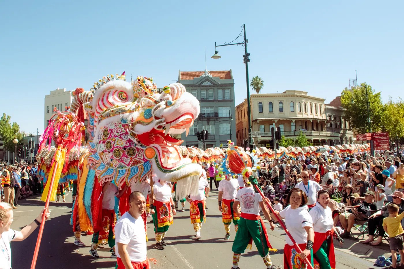A large dragon float, adorned with red, white, and yellow colors and featuring an elaborate headpiece, leads a parade down a city street lined with spectators and buildings.