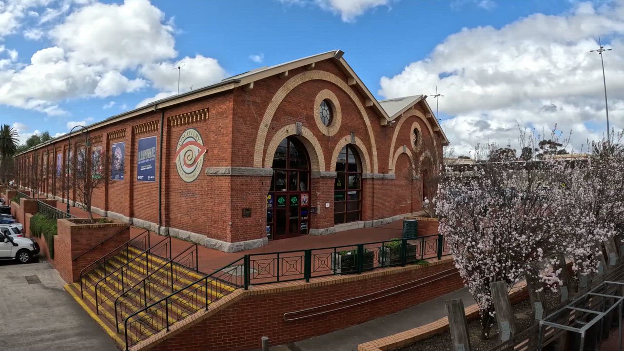 A red brick building with arched windows and decorative elements stands beside a paved area with trees and bushes under a blue sky with clouds.