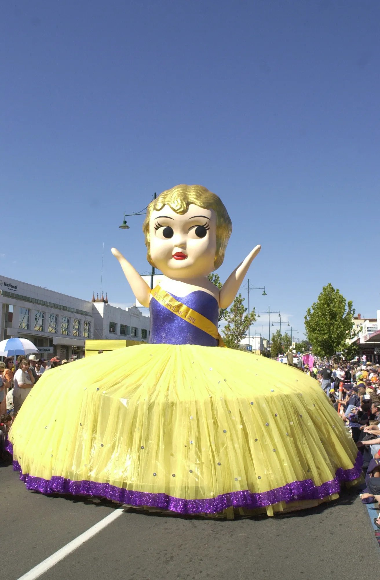 A large, yellow, doll-like figure with blonde hair and a purple sash floats down a street lined with spectators, under a clear blue sky.