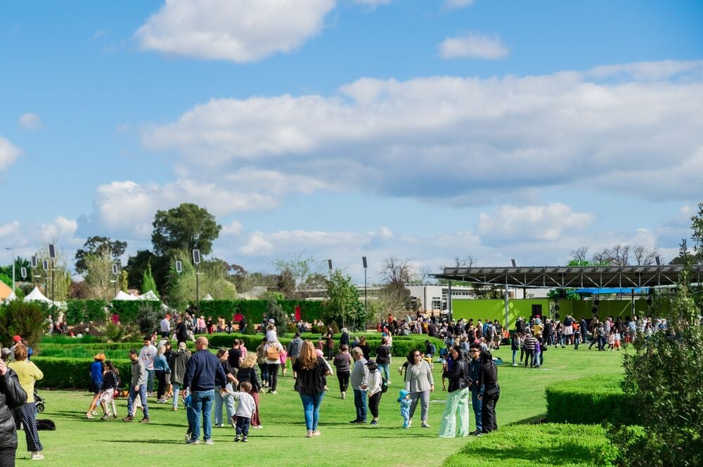 A large group of people gathers on a grassy lawn, with trees and a covered structure visible in the background.