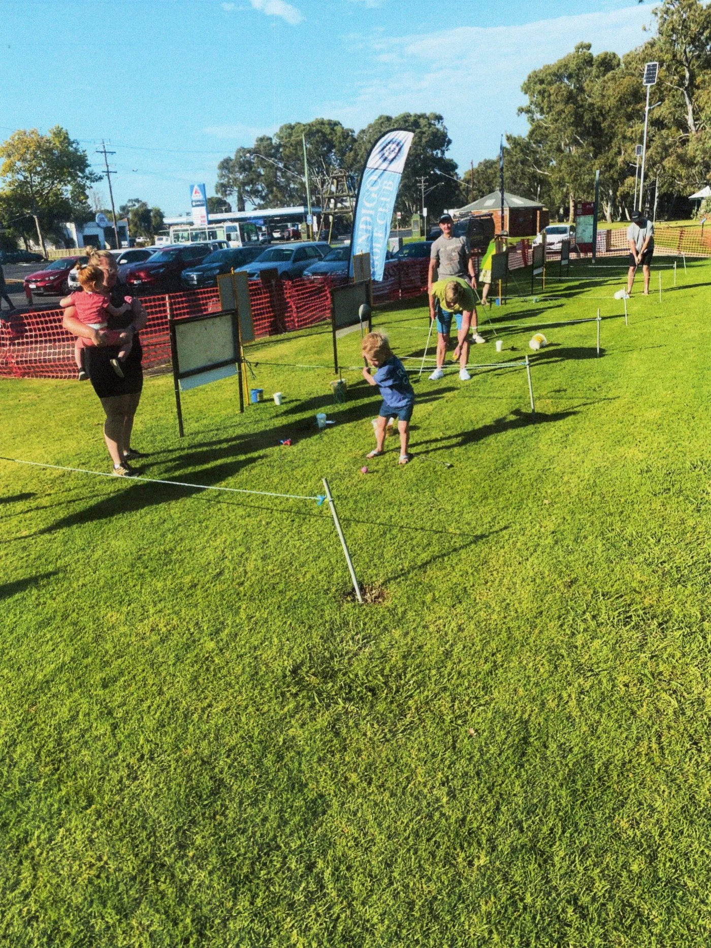 A child in a blue shirt stands on a grassy field, holding a small object, with a few adults nearby and a red fence and parking lot in the background.