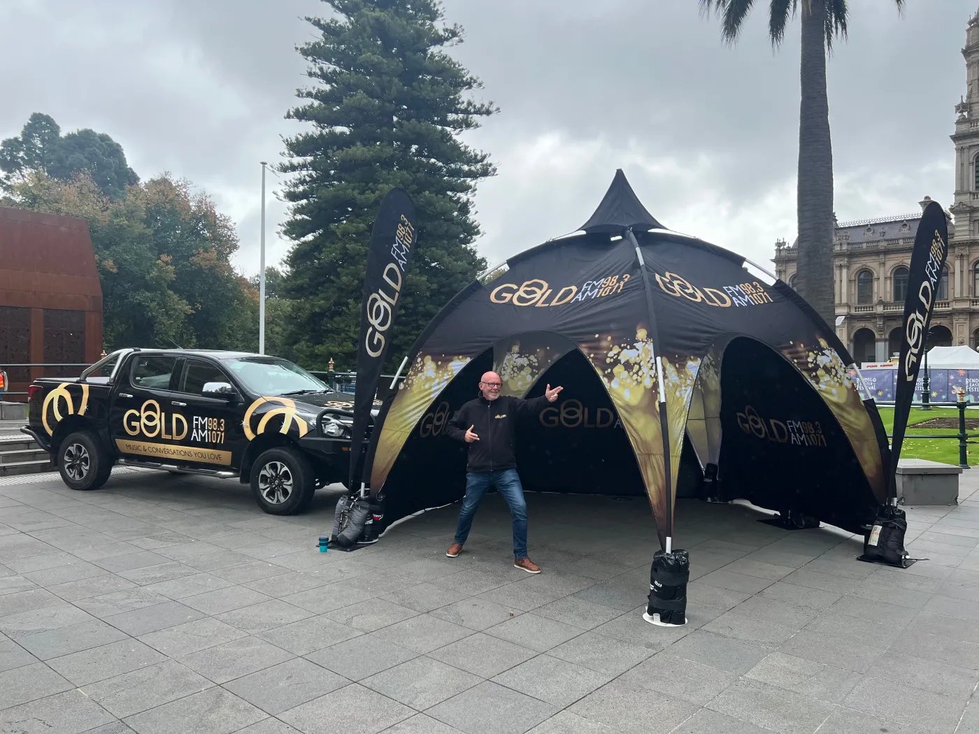 A man in a black shirt and blue jeans stands in front of a black tent with “GOLD” text and graphics, parked next to a black SUV with “GOLD” branding, in