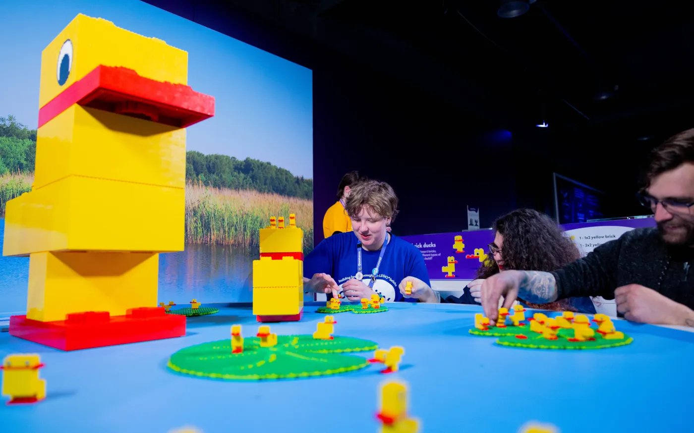 A large yellow Lego duck structure sits on a blue table with green circular bases, surrounded by small yellow Lego ducks, with three people nearby.