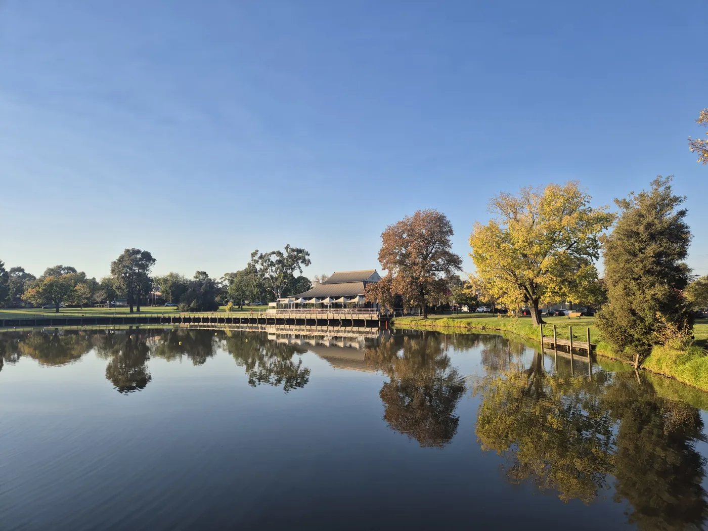A building with a brown roof sits on the edge of a calm body of water, with trees and grass visible on the opposite side, all under a clear blue sky.