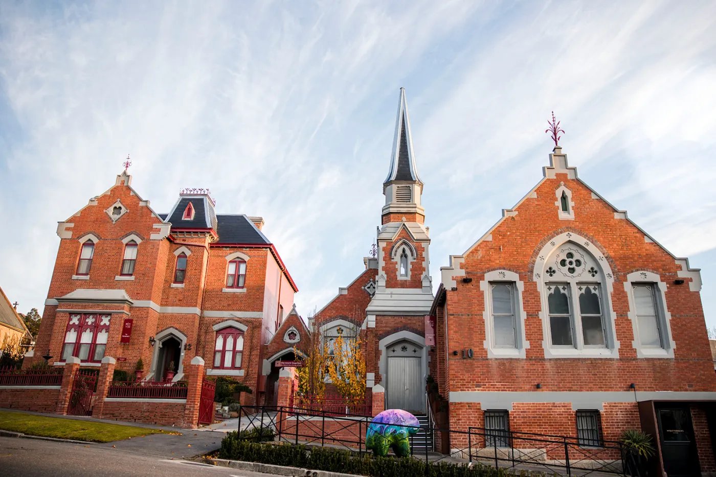 A red brick church with a tall steeple and cross stands beside a smaller red brick building under a partly cloudy sky, with a purple umbrella and fence in the foreground.