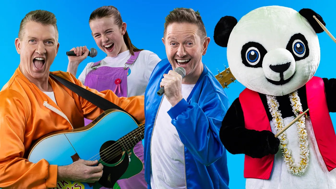 Four people stand against a blue background: one in an orange suit holds a guitar, another in a blue suit holds a microphone, a third in a purple outfit holds a microphone, and a panda
