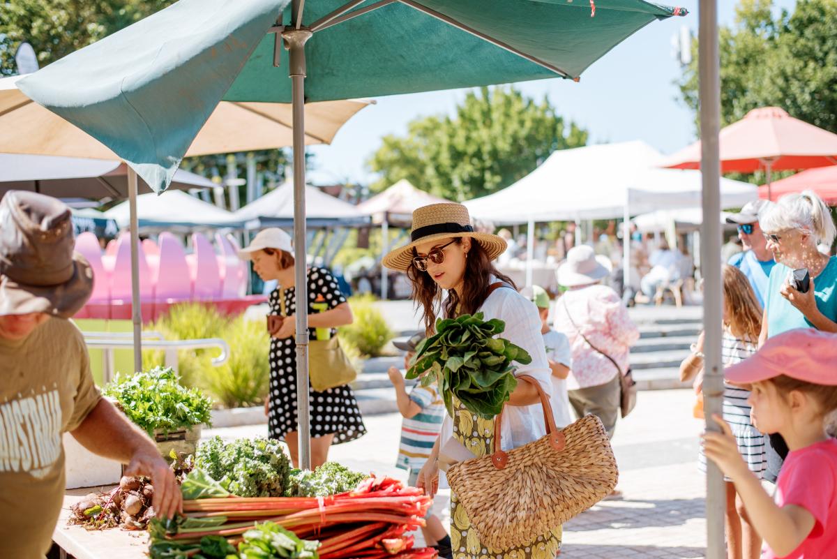 A woman in a white shirt and straw hat holds a basket of green leaves while shopping at a farmer's market under a green umbrella, with other market stalls and people in the background.