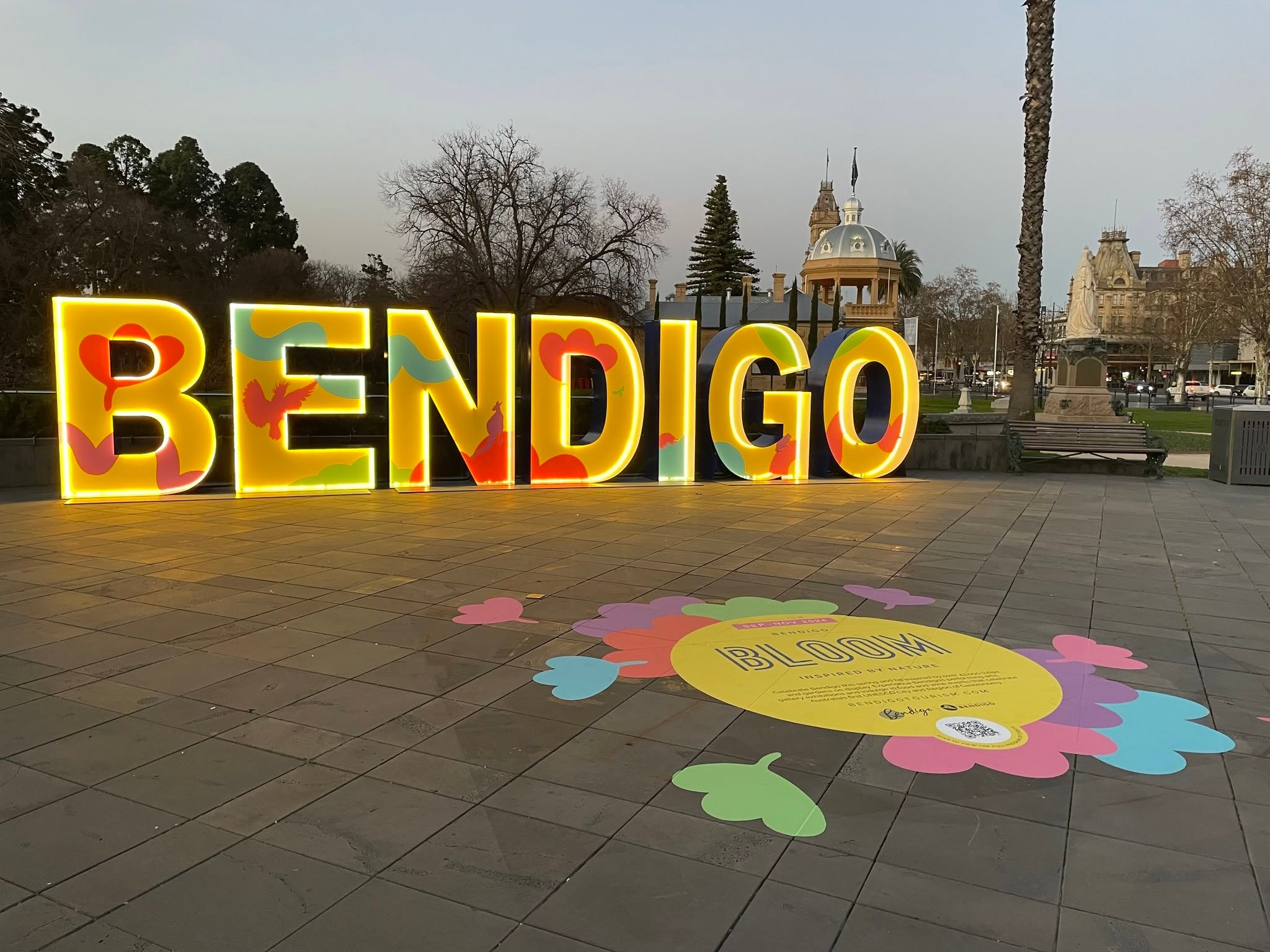 A large illuminated sign reading "BENDIGO" in yellow letters with colorful accents stands on a paved area, with colorful flower designs on the ground nearby and a building and trees in the background.