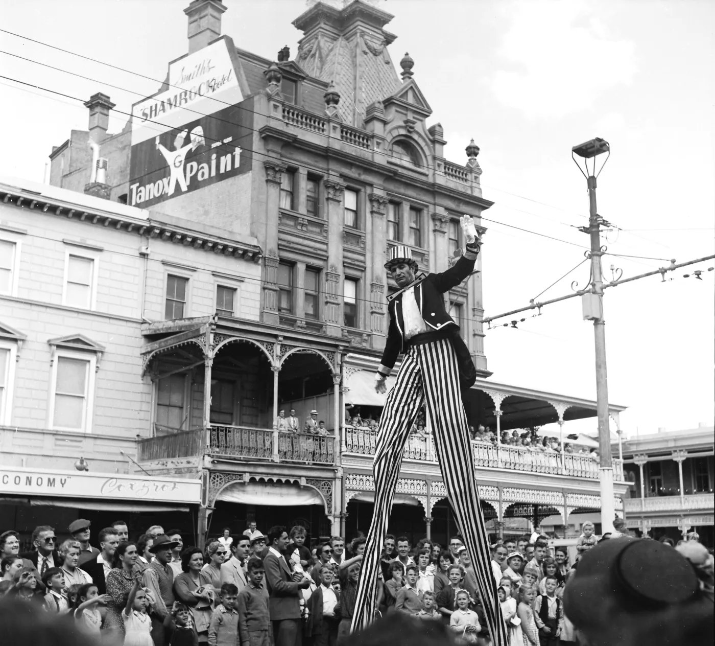 A man in a suit and striped pants stands on stilts in front of a large building, facing a crowd of onlookers. A sign for "Tanox Paint