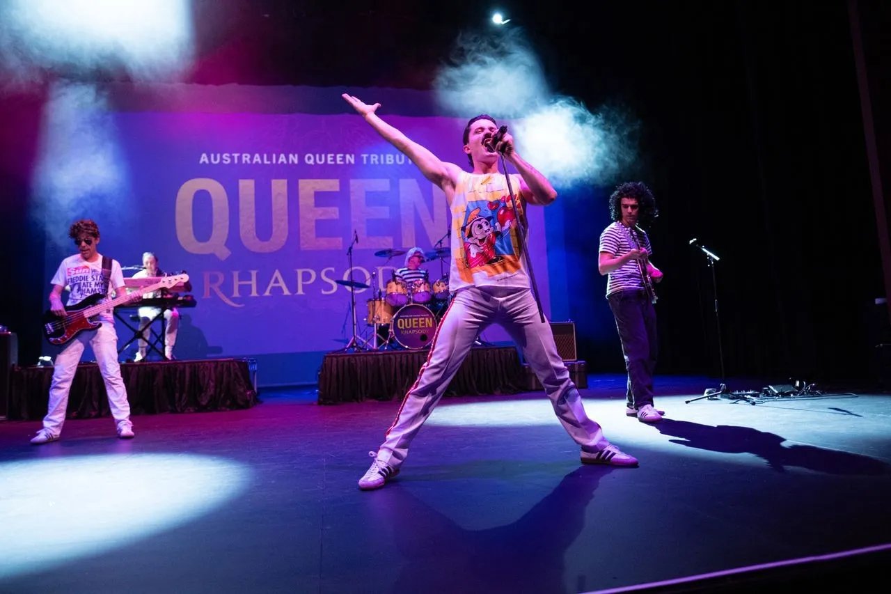 A band performs on a stage with a large “QUEEN RHAPSODY” banner and smoke effects. A singer in a yellow shirt with Minnie Mouse