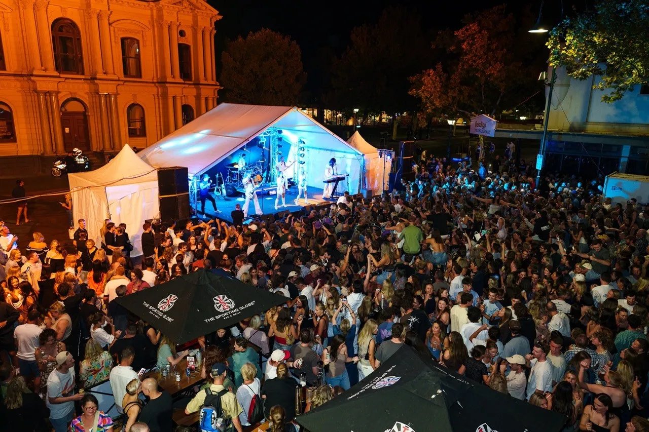 A large crowd gathers outside a tent-lit stage at night, with tents and illuminated buildings visible in the background.
