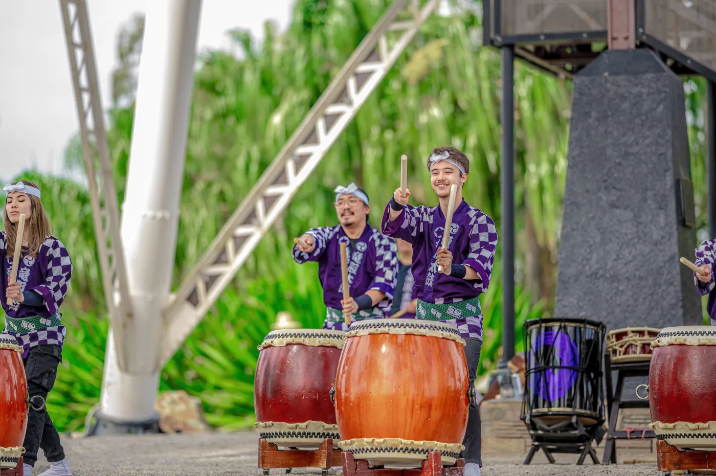 Four people in purple and white checkered shirts play large drums with wooden mallets on a paved surface, with a white metal structure and green foliage in the background.