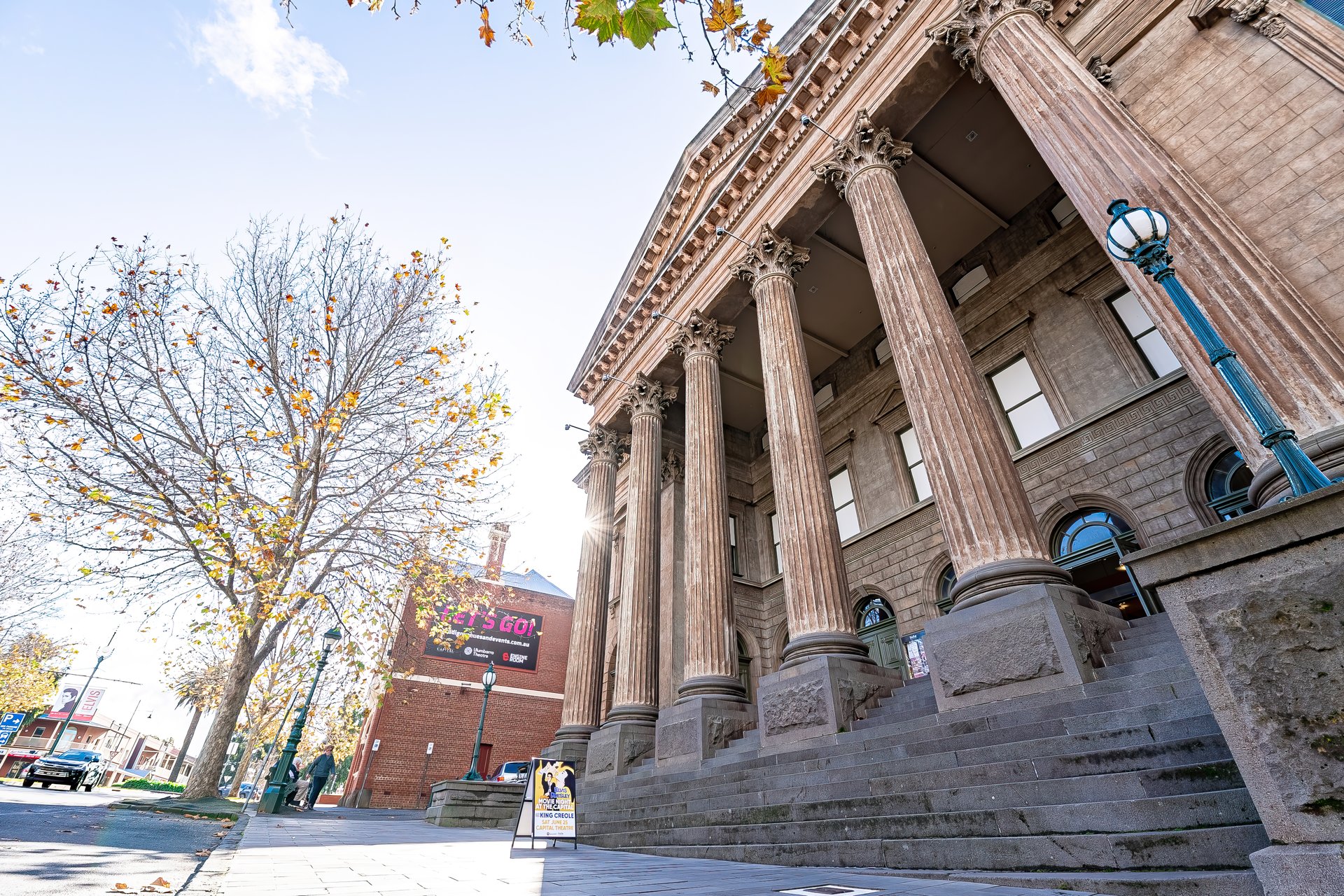 A grand stone building with classical columns and steps is pictured, with a tree and lamppost in the foreground, and a brick building visible behind it.