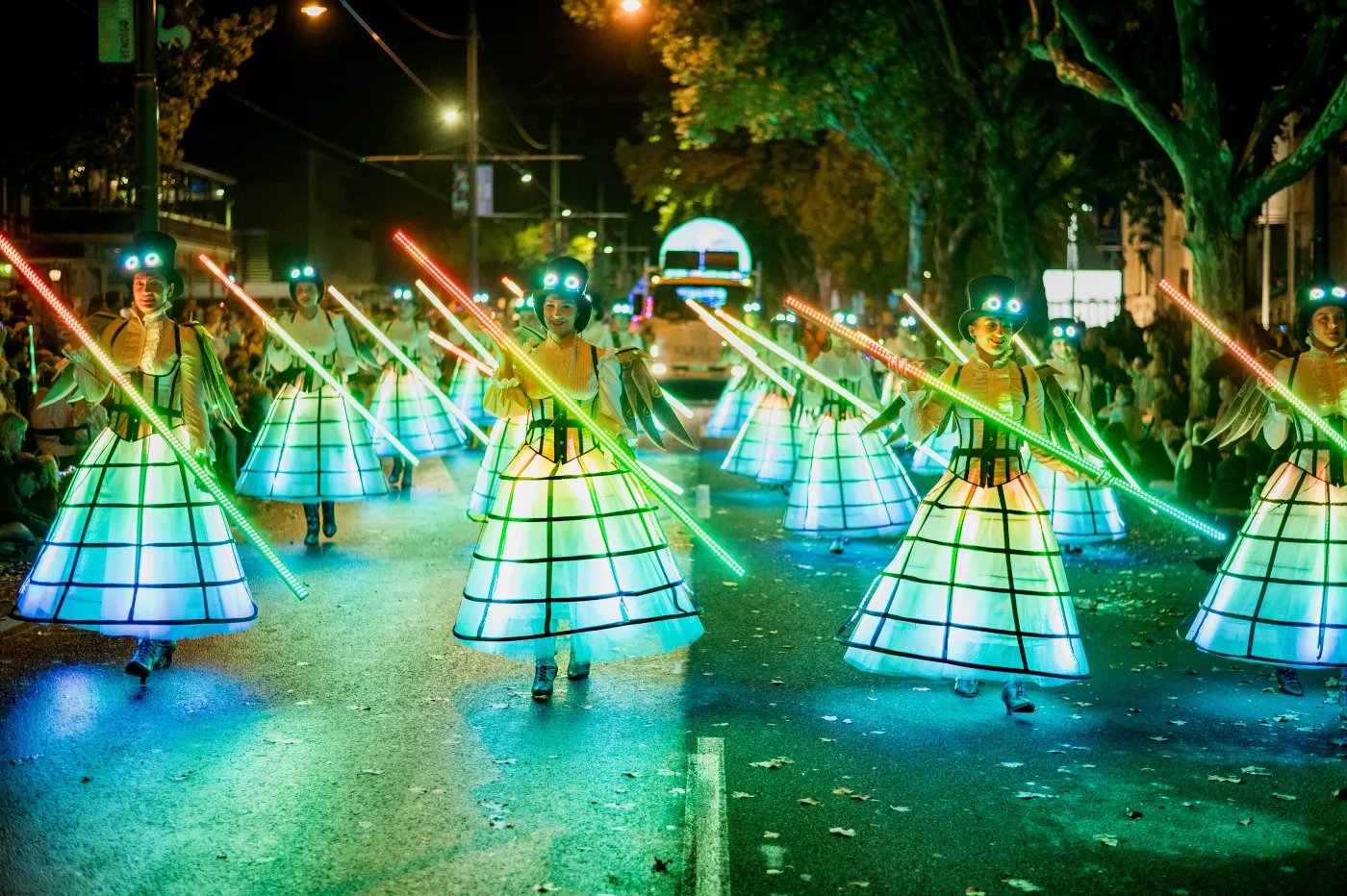 A group of performers in light-colored, grid-patterned dresses and headgear with glowing green and red accents march down a paved street at night, holding long green-lit swords.